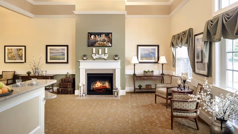 A cozy living room area with a lit fireplace in the center, flanked by framed artwork on the walls. There are two armchairs with patterned upholstery near a window with draped curtains, a small round table with decorative candles, and a console table with two lamps and plants. The room has beige walls, a carpeted floor, and a countertop with fruit and glasses on the left side.