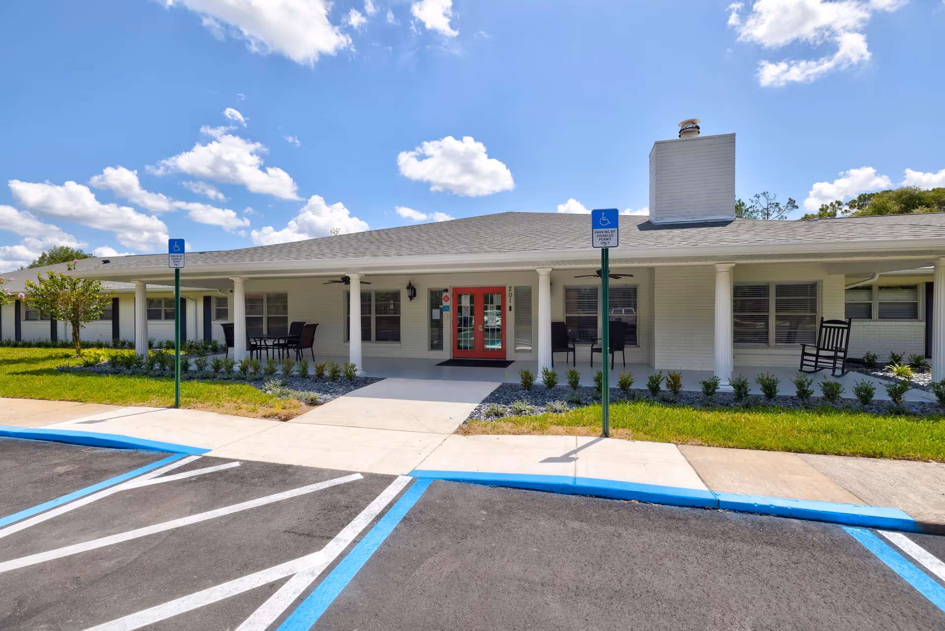 Exterior view of The Gardens of Eastbrooke facility showing a single-story building with white brick walls, a gray shingled roof, and a covered porch supported by white columns. The porch has outdoor seating including chairs and a table. There are two handicap parking spaces in front of the building with blue markings and signs. The sky is blue with scattered clouds.