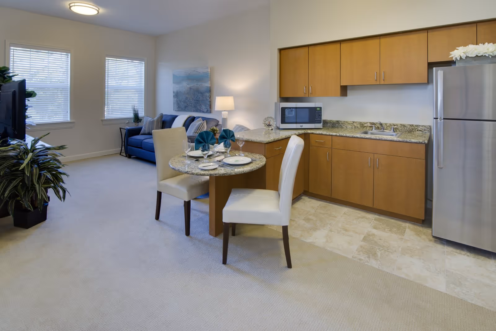 A bright and clean living space featuring a small kitchen area with wooden cabinets, a granite countertop, a microwave, and a stainless steel refrigerator. Adjacent to the kitchen is a round dining table set for two with white chairs and teal napkins. In the background, there is a blue sofa with cushions, a side table with a lamp, and two windows with blinds. A large green plant is visible near a television on the left side.