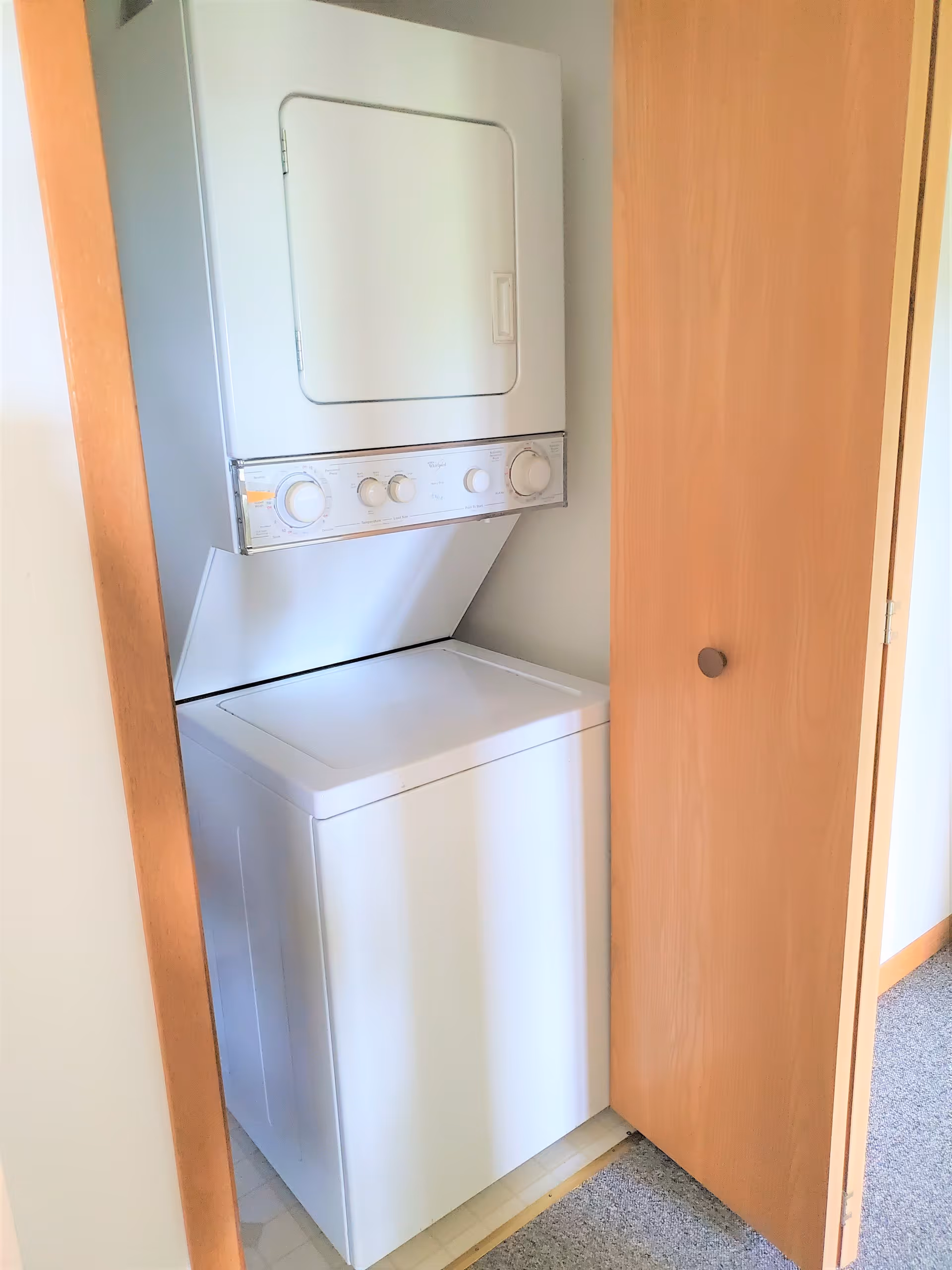 A compact stacked washer and dryer unit inside a small closet with wooden bi-fold doors, located in a residential setting with carpeted flooring outside the closet.