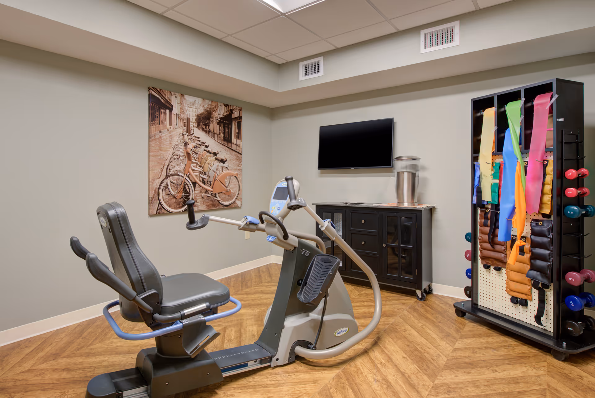 A small exercise room with a recumbent exercise bike in the center. On the right side, there is a rack holding colorful resistance bands and various dumbbells. A black cabinet with glass doors is against the wall, topped with a water dispenser. A flat-screen TV is mounted on the wall above the cabinet. A large framed photo of bicycles lined up on a street hangs on the left wall. The room has wood-patterned flooring and light-colored walls.