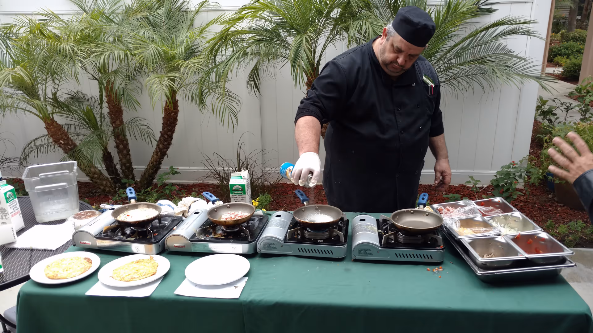 A chef dressed in black is cooking outdoors on four portable gas stoves placed on a green tablecloth-covered table. Various ingredients and cooking utensils are arranged on the table, including plates with cooked food, cartons of milk, and metal trays with chopped vegetables. The background features a white fence and several palm plants.