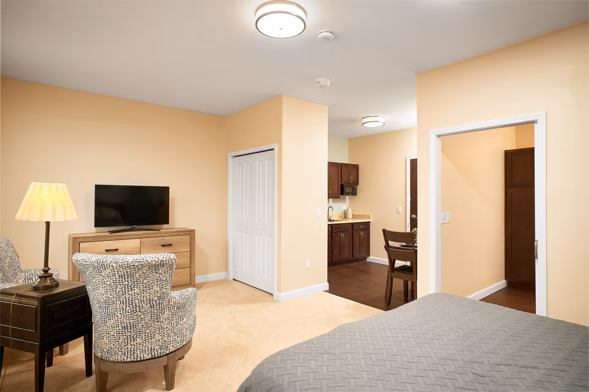Interior view of a senior living facility room with beige walls and carpet. The room features a bed with a gray quilt, two patterned armchairs, a wooden dresser with a flat-screen TV on top, a side table with a lamp, and an open doorway leading to a kitchen area with dark wood cabinets and a dining table with chairs.