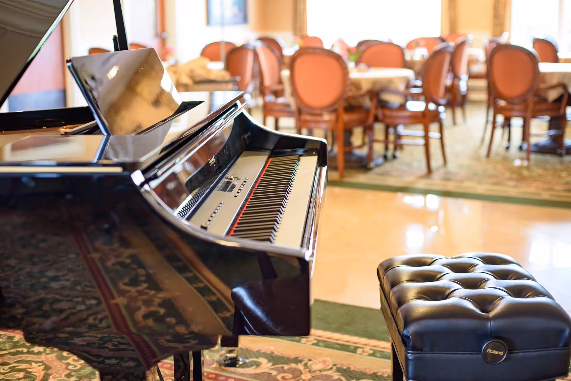 Close-up view of a black grand piano with a padded piano bench in the foreground, set in a room with carpet and polished floors. In the background, there are round dining tables with chairs upholstered in a reddish-brown fabric.