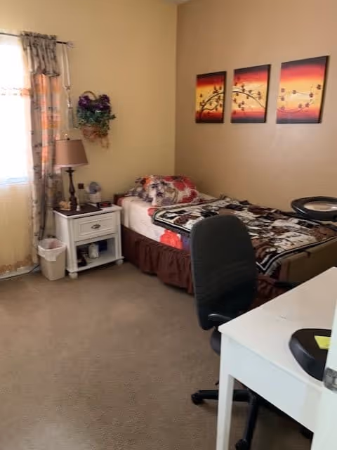 A cozy bedroom with a single bed covered in a floral blanket, a white nightstand with a lamp and some items on it, a window with sheer curtains, a black office chair, and a white desk. Three colorful paintings hang on the beige wall above the bed.