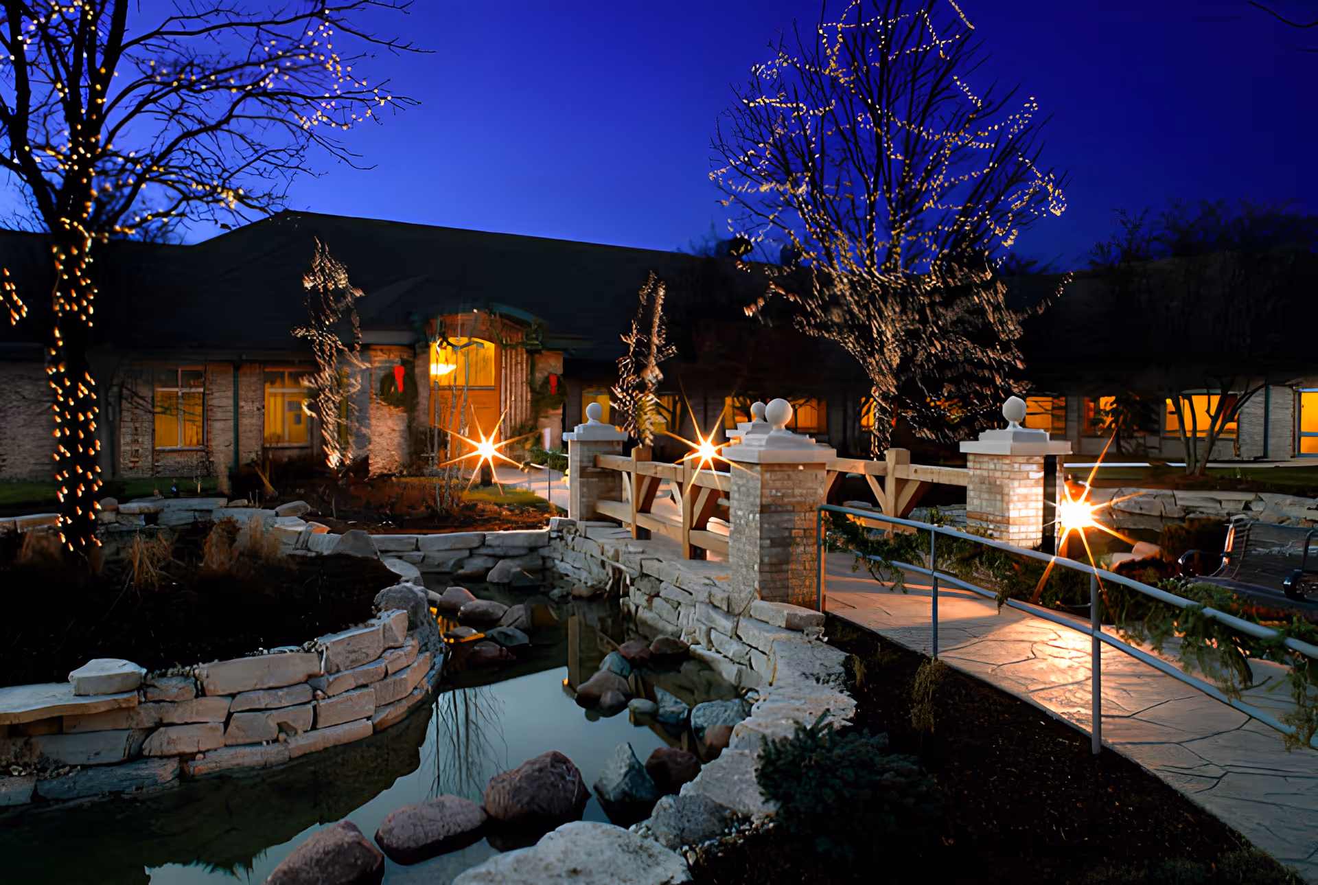 Evening view of a senior living facility entrance with a stone bridge over a small pond, trees decorated with string lights, and warm lights glowing from inside the building.
