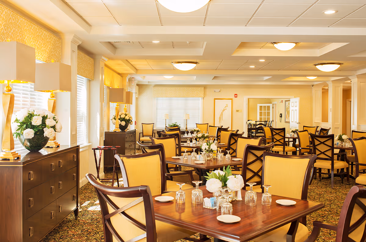 Sunlit, elegant dining room with wooden tables, yellow upholstered chairs, floral centerpieces, and sideboards with lamps.