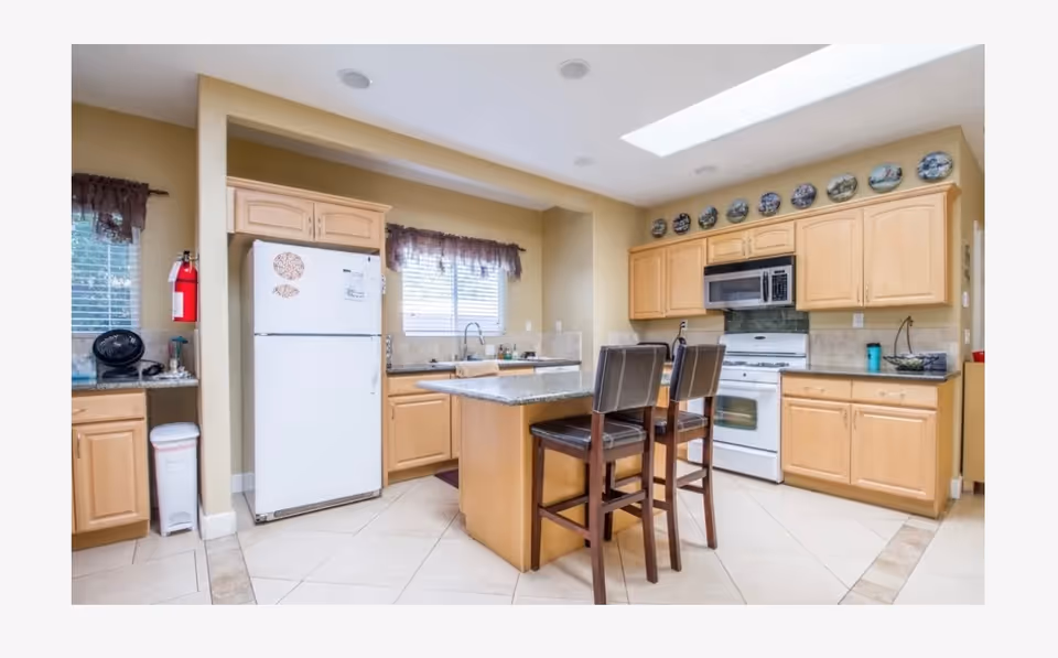 A bright kitchen with light wood cabinets, a white refrigerator, a white stove with a microwave above it, and a central island with a granite countertop. Two dark wooden chairs with black cushions are placed at the island. There are windows with brown curtains, a fire extinguisher mounted on the wall, and decorative plates displayed above the cabinets.