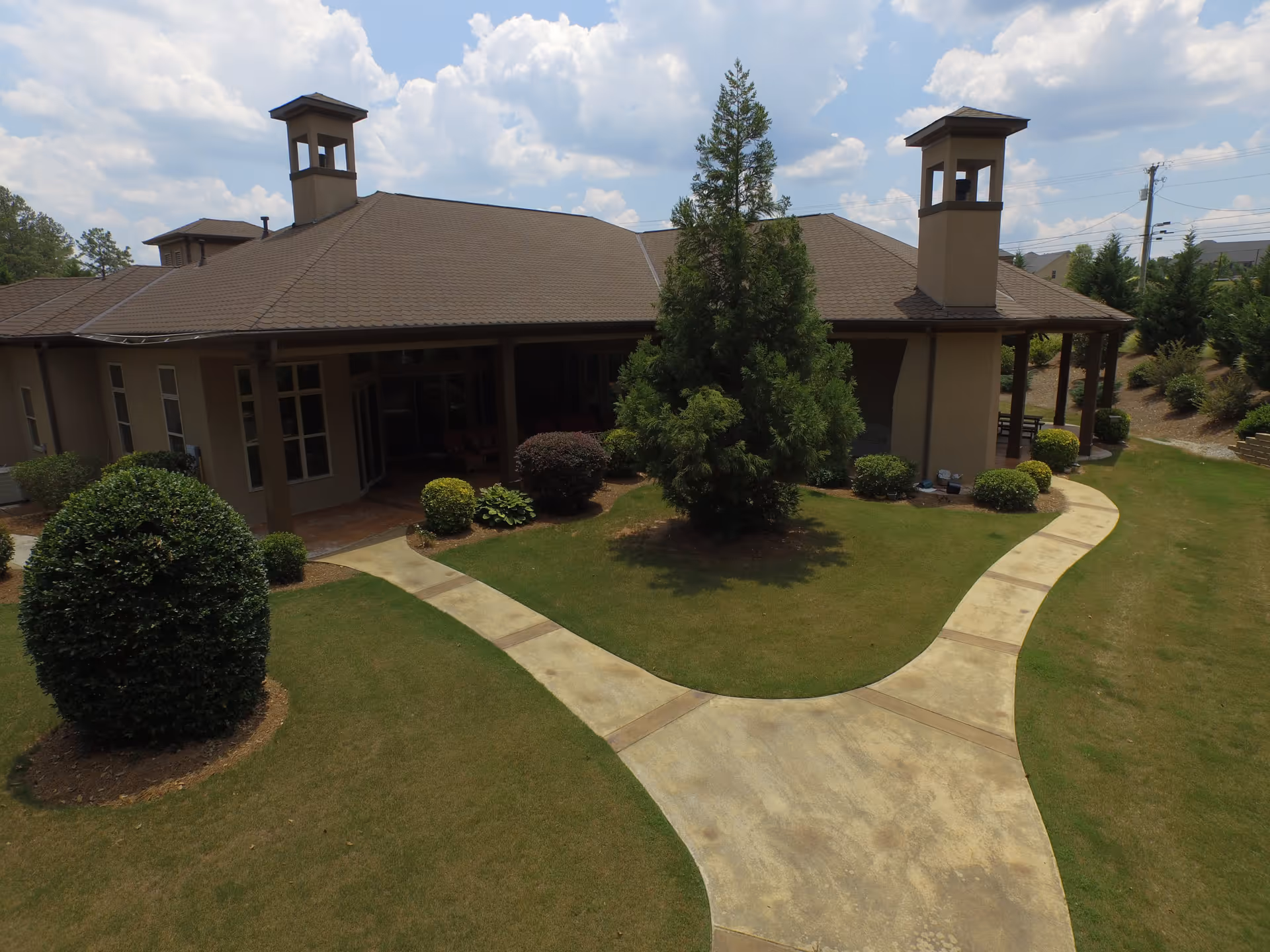 Exterior view of a single-story senior living facility building with a brown roof and beige walls, surrounded by well-maintained green lawns, bushes, and trees. A concrete pathway splits into two directions leading to the building entrance and around the side. The sky is partly cloudy.
