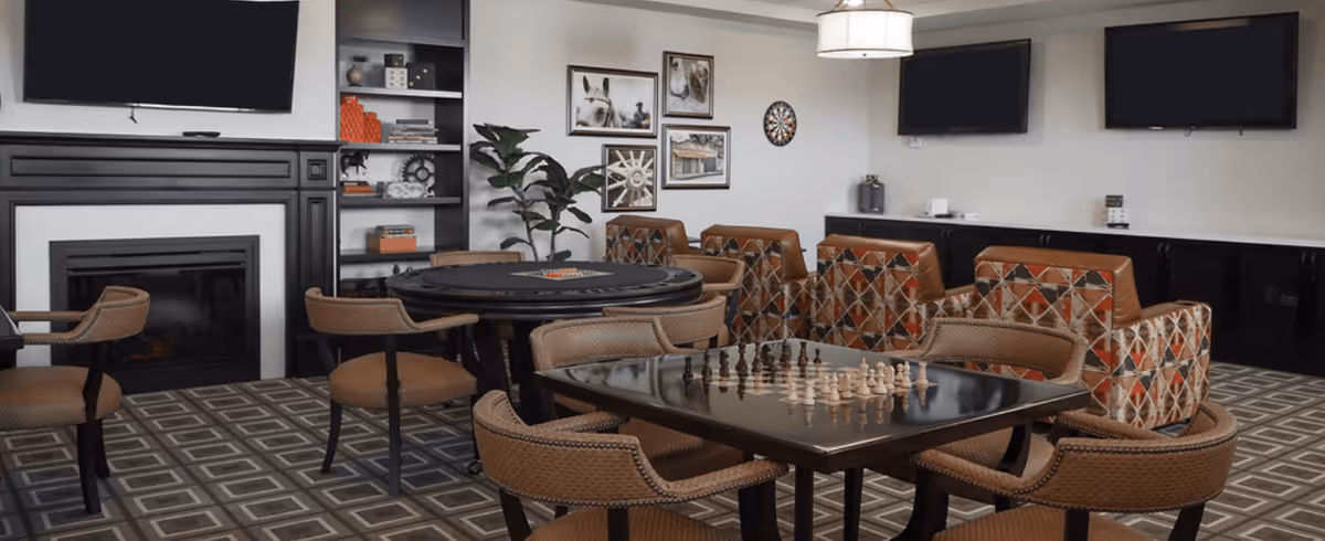 A cozy common area with a fireplace, two flat-screen TVs mounted on the walls, a chess table with chess pieces set up, a round table with a game on it, several cushioned chairs, patterned armchairs, a dartboard on the wall, and decorative framed pictures. The room has a patterned carpet and a modern light fixture hanging from the ceiling.