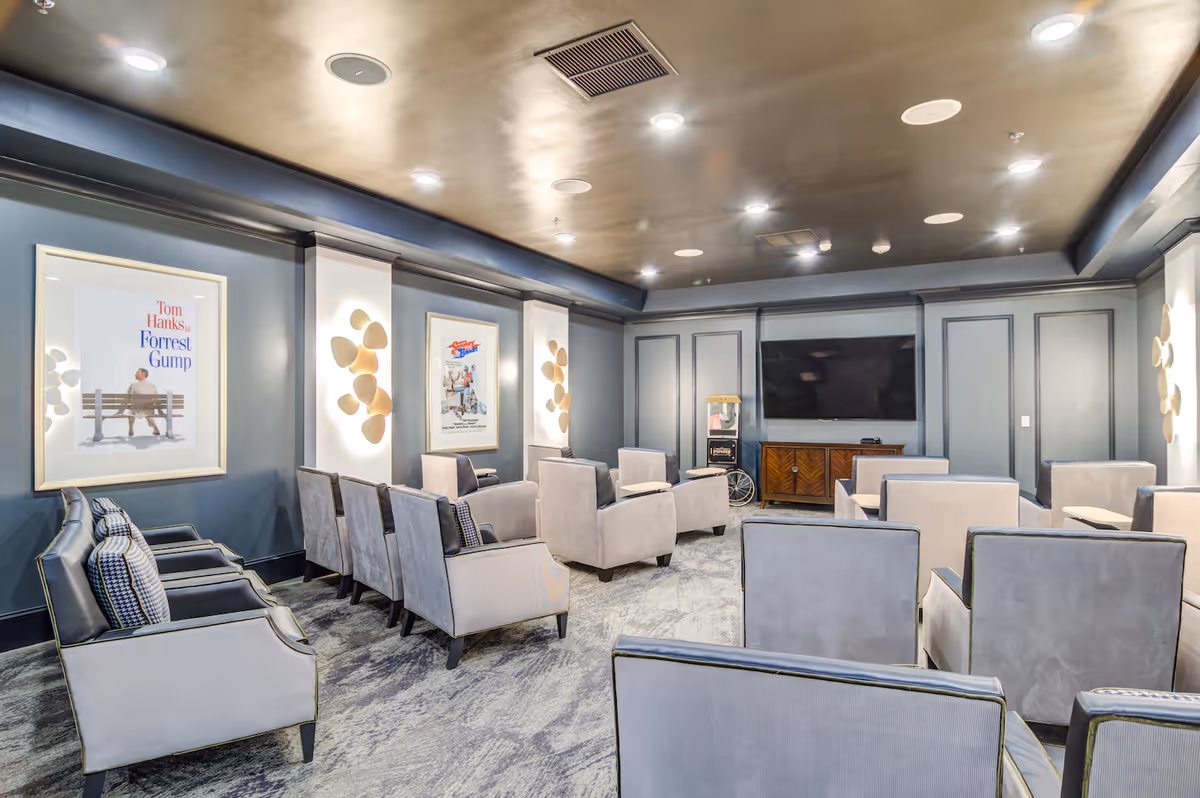 A community media room with rows of gray upholstered chairs facing a wall-mounted TV and framed movie posters on the walls.