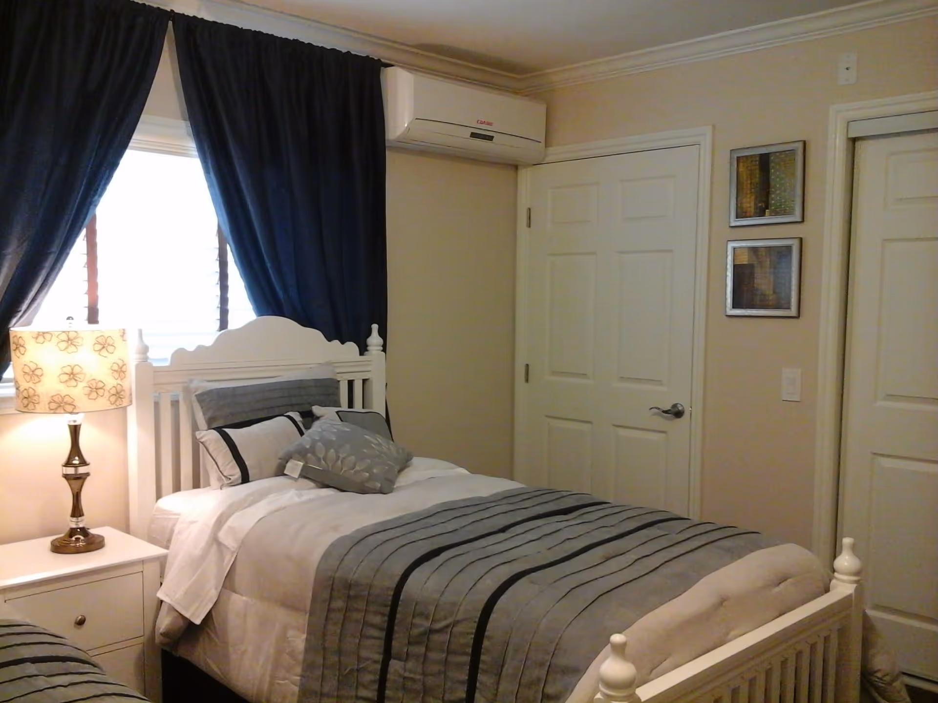 A bedroom with a white wooden bed frame and a neatly made bed with gray and white bedding. There is a nightstand with a lamp featuring a floral patterned shade next to the bed. Dark curtains cover a window behind the bed. On the wall, there is a white air conditioning unit above a closed door, and two framed pictures hang on the adjacent wall.
