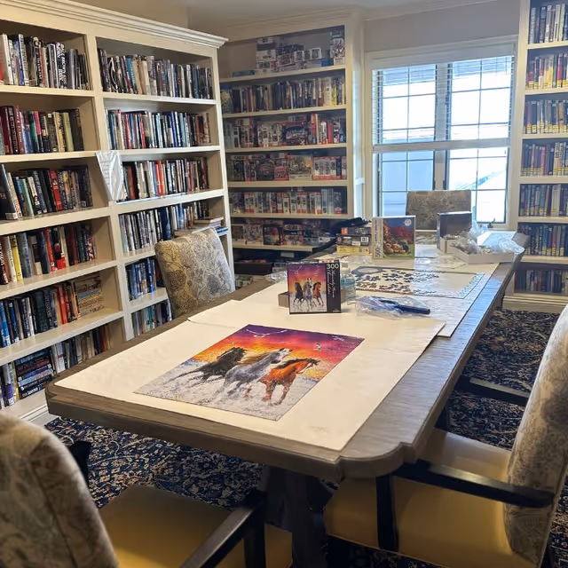 A cozy room with bookshelves filled with books and puzzles along the walls. A large table in the center has a partially completed horse-themed jigsaw puzzle and several puzzle boxes on it. Four upholstered chairs surround the table, and a window lets in natural light.
