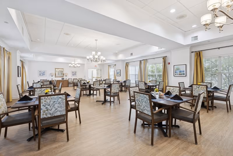 A spacious dining room with multiple wooden tables and chairs arranged neatly. Each table has black napkins and some have small flower arrangements. The room has large windows with yellow curtains, light wood flooring, white walls, and ceiling lights including chandeliers. Artwork is hung on the walls, and the room is brightly lit with natural and artificial light.