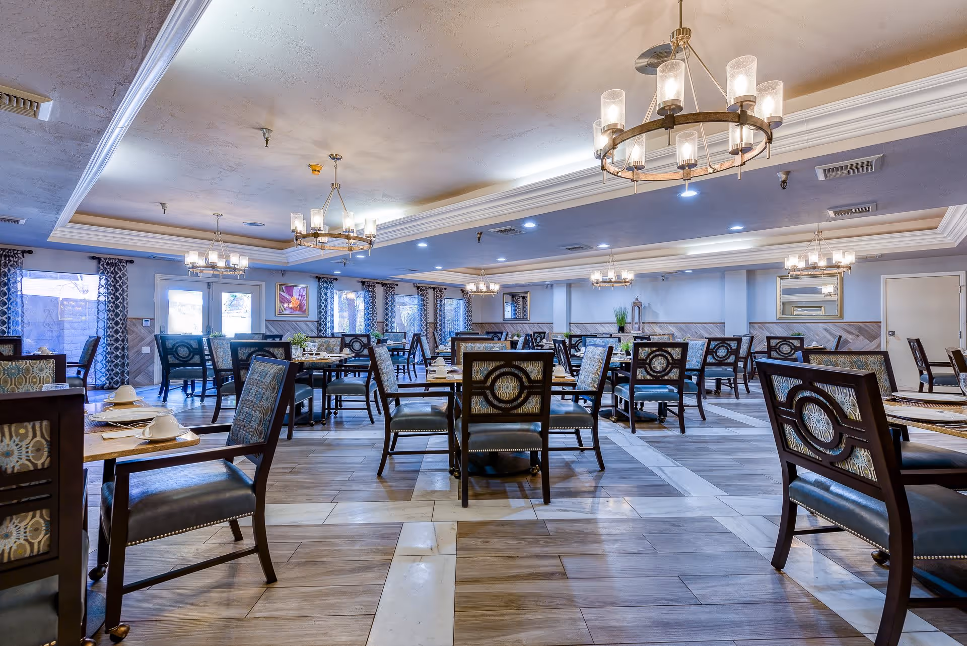 Spacious dining room with multiple wooden tables and chairs arranged neatly. The room features large windows with patterned curtains, elegant chandeliers hanging from a coffered ceiling, and a mix of light wood and tile flooring. The walls are painted in soft tones with decorative artwork and mirrors.