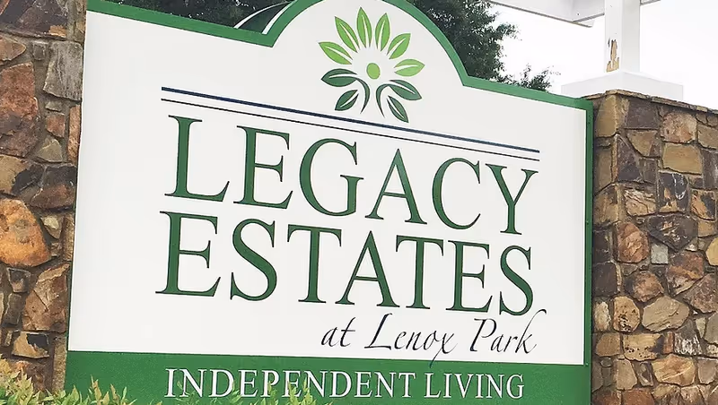 A large outdoor sign for Legacy Estates at Lenox Park, an independent living facility, mounted on a stone wall with a green and white color scheme and a green floral logo above the text.