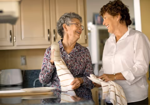Two elderly women smiling and drying dishes together in a kitchen with light-colored cabinets and a toaster visible on the counter.