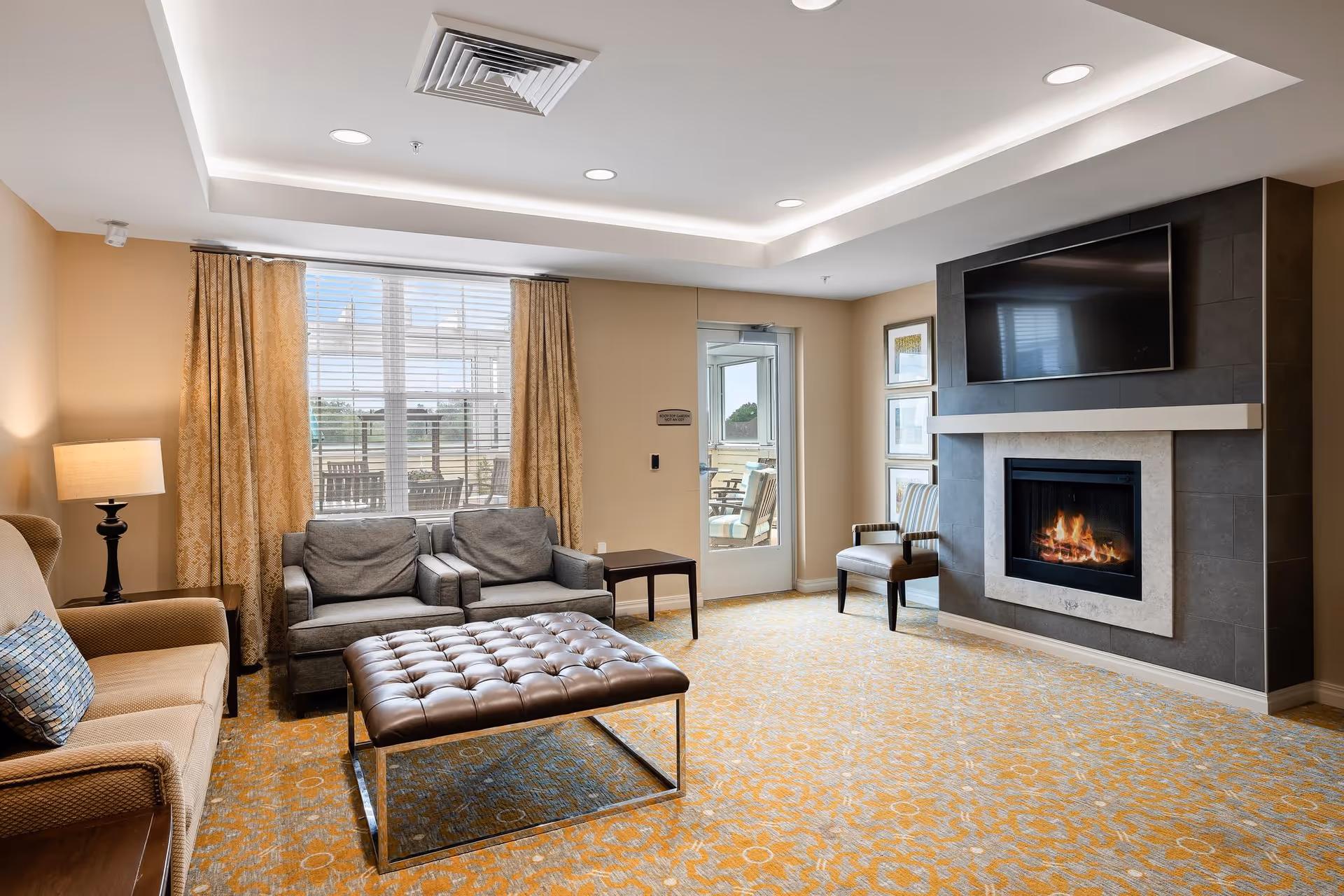 Lounge area with sofas, a large tufted ottoman, a fireplace with a TV above, and a window letting in natural light.