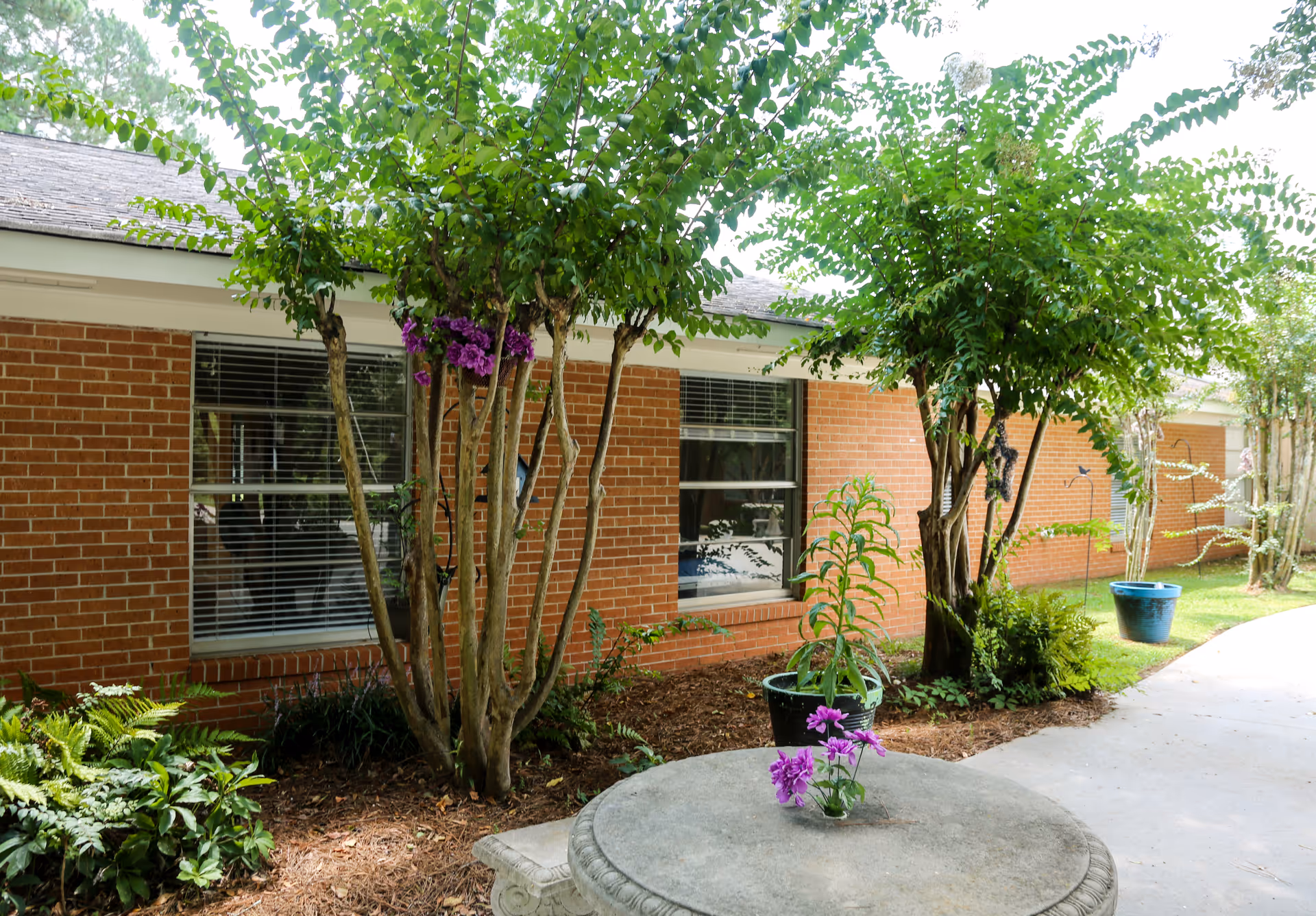 Outdoor garden area at Eatonton Health & Rehabilitation featuring a brick building with windows, several trees and plants, a concrete round table with a small vase of purple flowers, and a paved walkway.