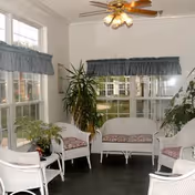 Bright sunroom with white wicker chairs and a loveseat arranged around potted plants beneath a ceiling fan.