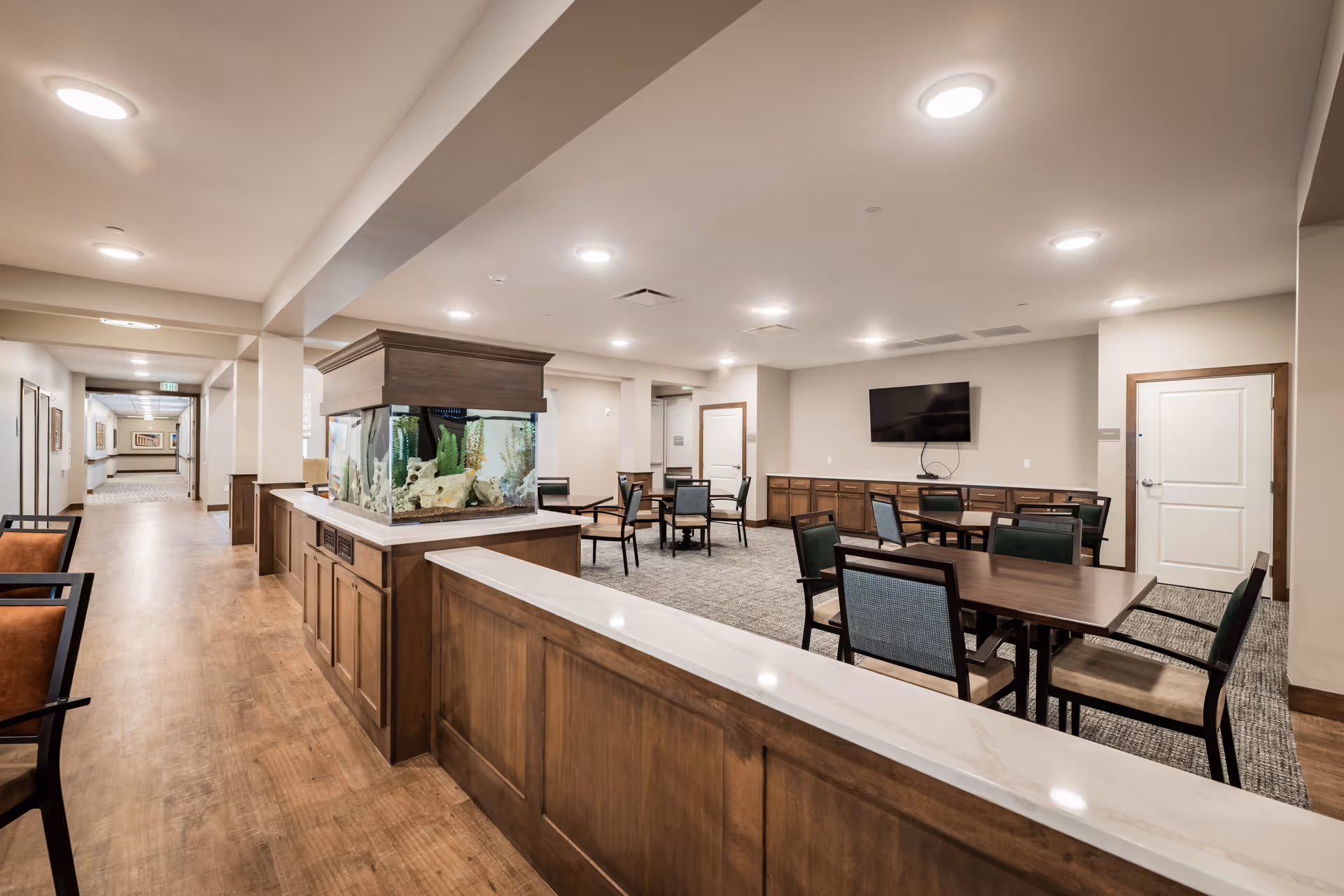 Interior view of a senior living facility common area with wooden flooring, a large fish tank on a wooden cabinet, several tables and chairs, a wall-mounted TV, and bright ceiling lights.