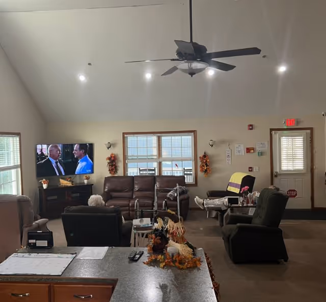 Communal living room with sofas and recliners facing a TV, a ceiling fan, and fall decorations.