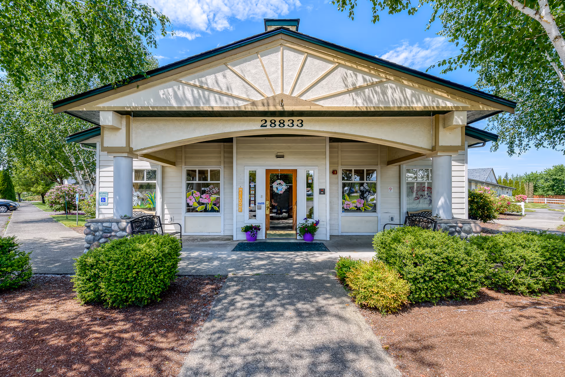 Front exterior view of a single-story assisted living facility building with the address number 28833 displayed above the entrance. The entrance has a covered porch supported by columns with stone bases, and there are decorative flower paintings on the windows on either side of the door. The surrounding area includes green bushes, trees, and a clear blue sky.