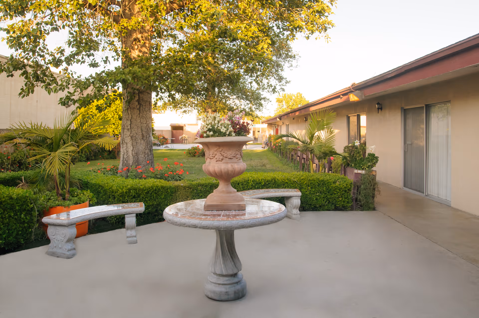 Outdoor patio area at Orangegrove Rehabilitation Hospital featuring a round stone table with a large decorative planter filled with flowers on top, two curved stone benches, neatly trimmed hedges, potted plants, a large tree, and a building with sliding glass doors in the background.