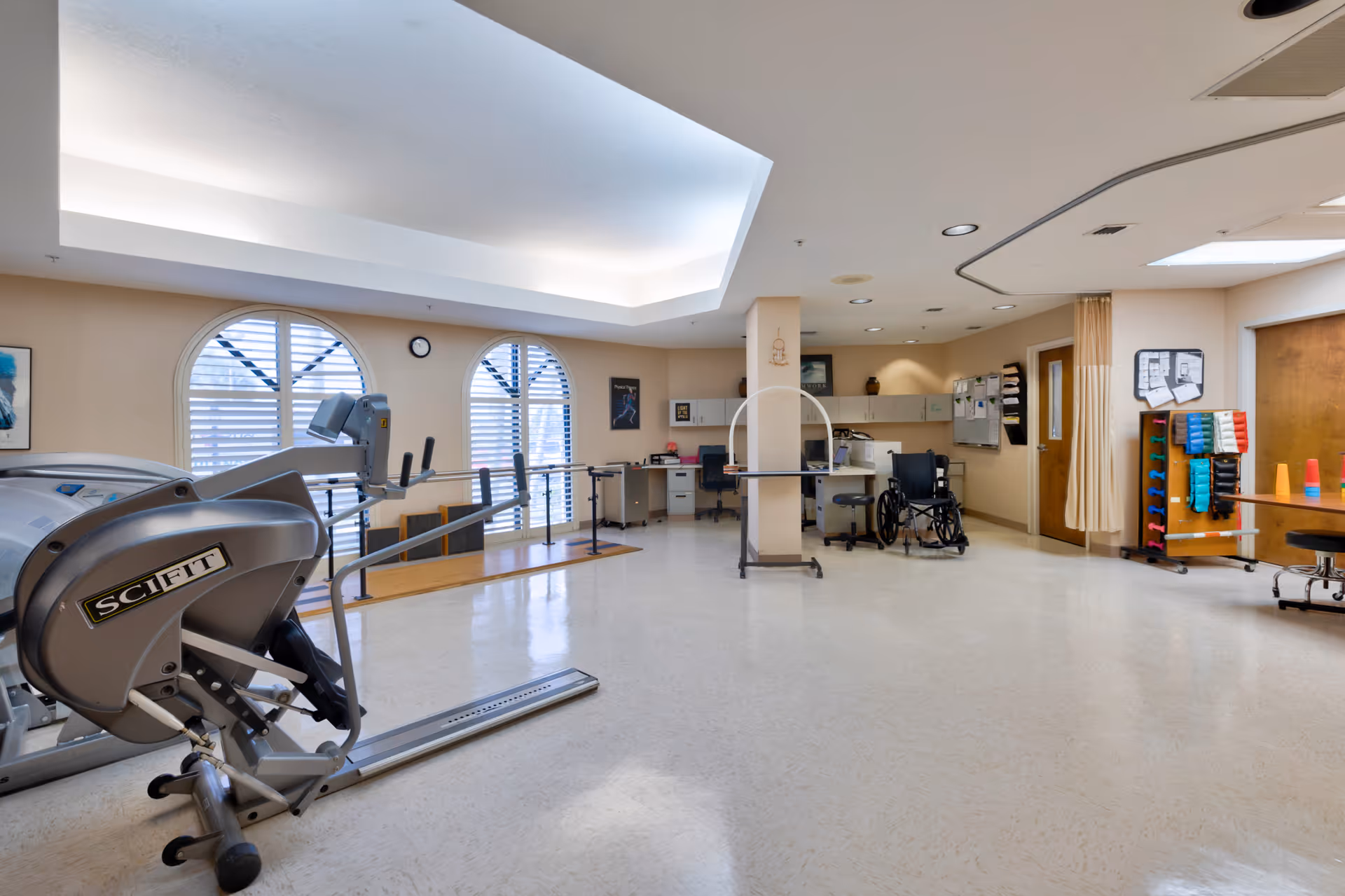 A spacious rehabilitation or physical therapy room with exercise equipment including a SciFit machine, parallel bars for walking practice, a wheelchair, desks with office chairs, and shelves with colorful exercise bands and cones. The room has large arched windows with white shutters and a bright ceiling light.