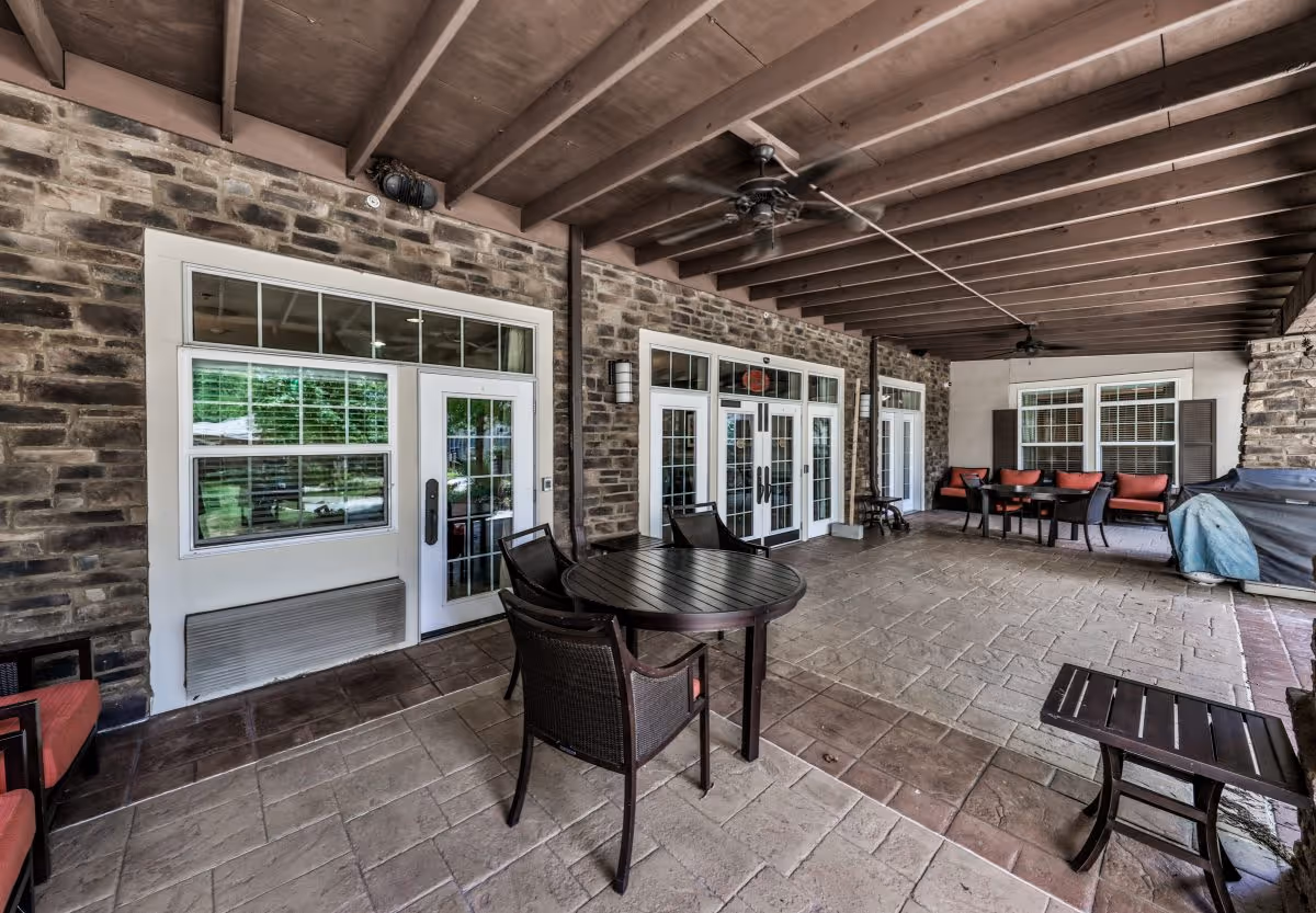 Covered outdoor patio area with stone walls and ceiling beams, featuring multiple seating arrangements including round tables with chairs and cushioned benches. Ceiling fans are mounted on the wooden beam ceiling, and there is a grill covered with a blue tarp on the right side.