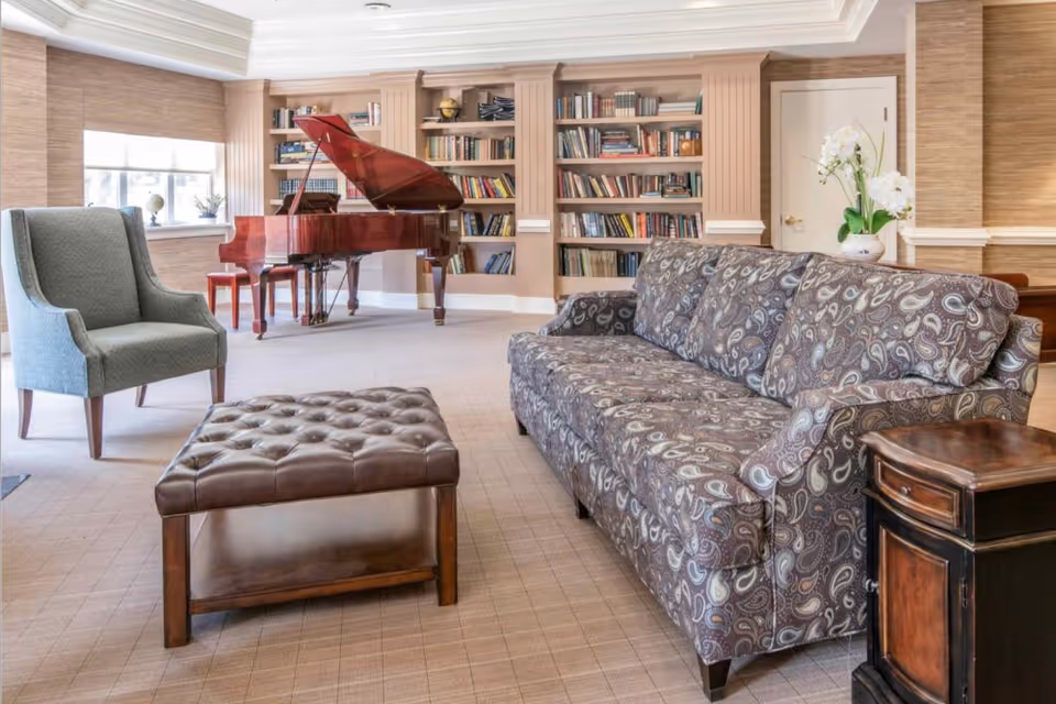 Bright communal living area with a patterned sofa, armchair, tufted leather ottoman, grand piano, and built-in bookshelves.