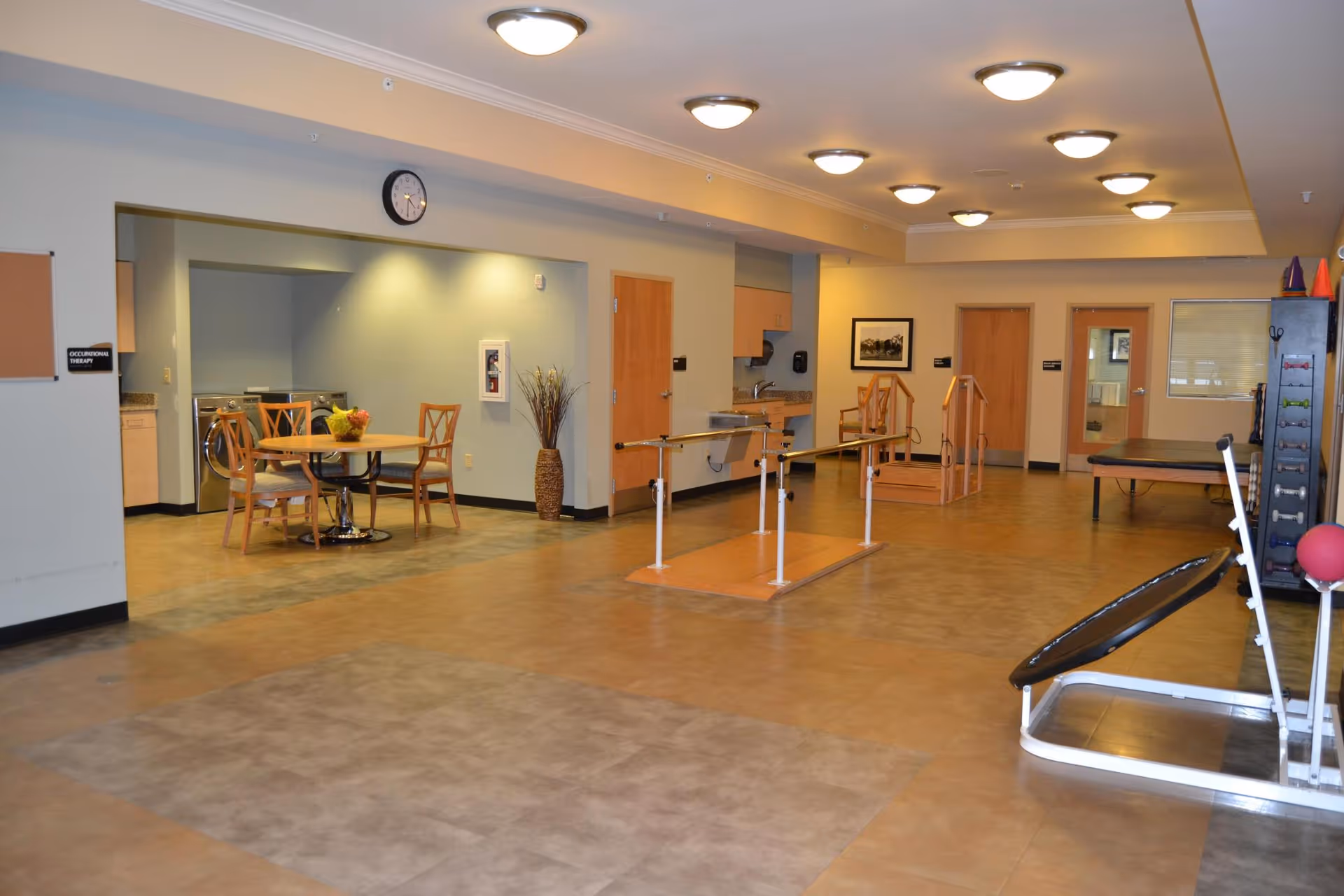 A spacious rehabilitation therapy room with wooden parallel bars for walking exercises, a small round table with four chairs, exercise equipment including a rebounder and weights, and a therapy table. The room has beige walls, multiple ceiling lights, and several doors leading to other areas. A clock is mounted above the table area, and a sign on the wall reads 'Occupational Therapy'.