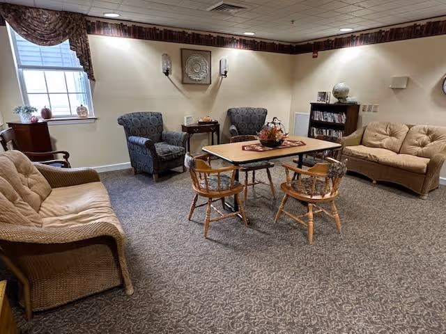 Carpeted common lounge with sofas, armchairs, a central table with wooden chairs, a bookshelf and a window.