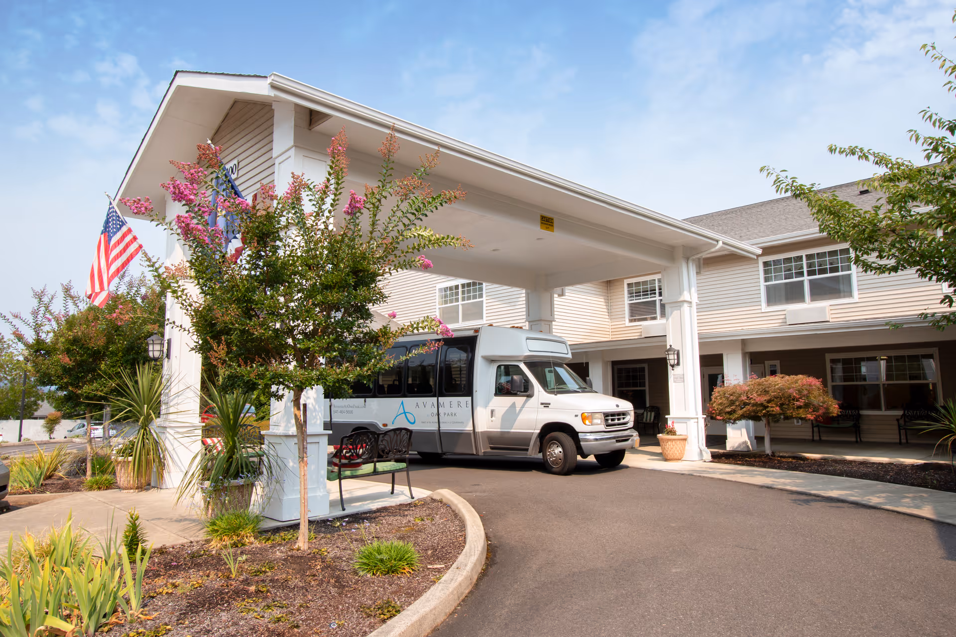 Entrance of a senior living facility with a covered drop-off area. A white shuttle van with the logo 'Avamere Oak Park' is parked under the canopy. There are small trees, plants, and an American flag near the entrance. The building is two stories with beige siding and multiple windows.