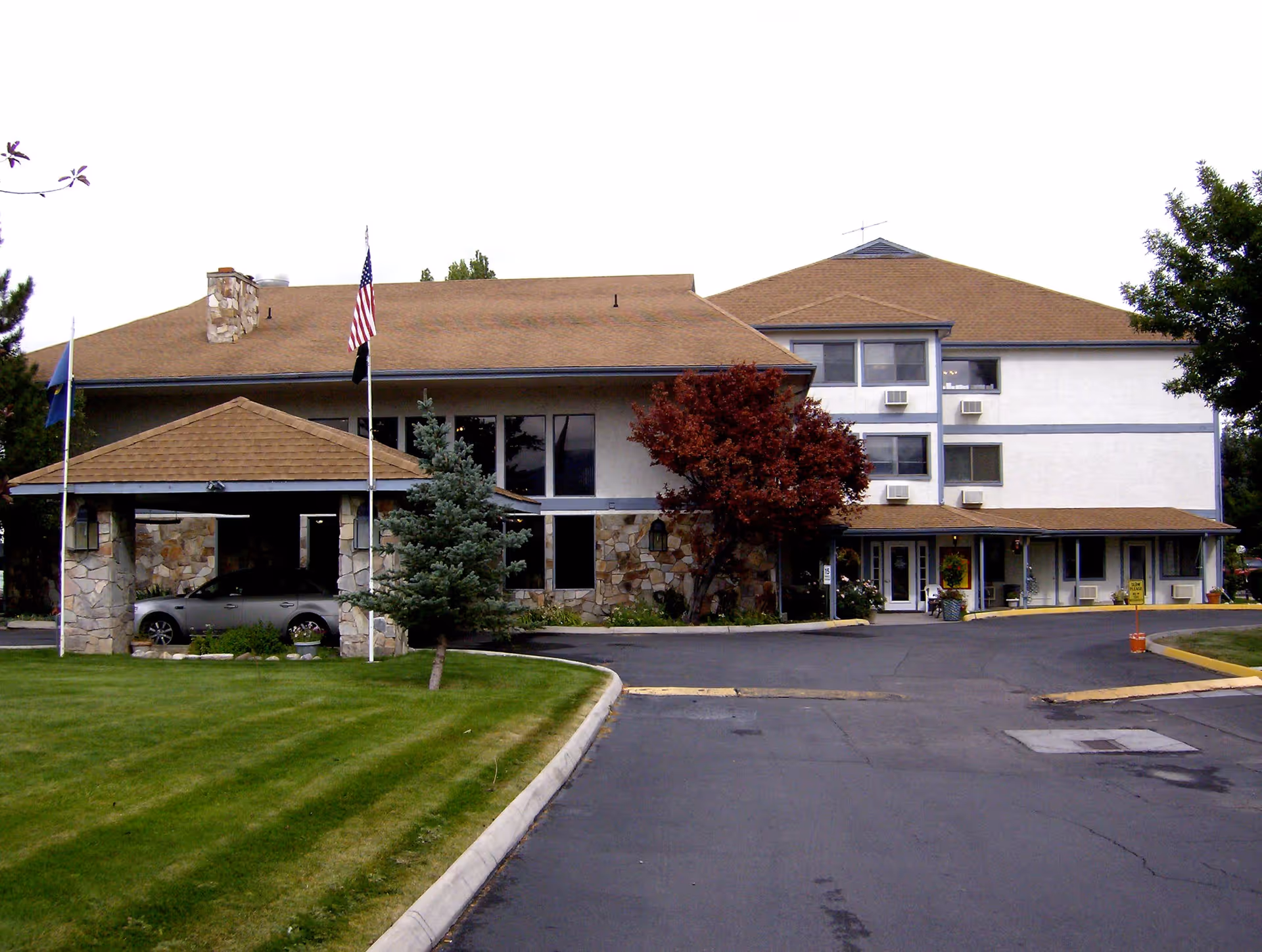 Exterior front view of a multi-story retirement community building with a covered entrance, stone and stucco facade, several windows with air conditioning units, a driveway, and well-maintained lawn with trees and flagpoles displaying the American flag and another flag.