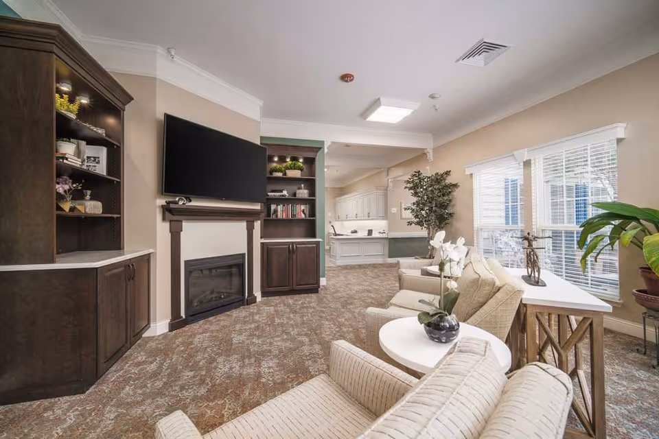 A cozy living room area in a senior living facility with beige walls and carpet. The room features a mounted flat-screen TV above a fireplace, built-in dark wood shelves with decorative items and books, and several beige upholstered armchairs arranged around small white tables with potted plants. Large windows with white blinds allow natural light to fill the space, and there is a green plant near the window. The room opens into a hallway with white cabinetry and a countertop.