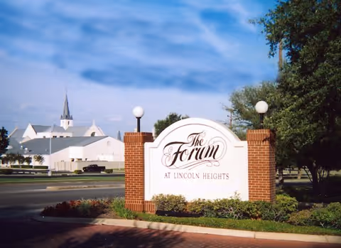 A large white sign with brick pillars on each side displaying the text 'The Forum at Lincoln Heights' situated on a landscaped area near a road. In the background, there is a white building and a church with a tall steeple under a partly cloudy blue sky.