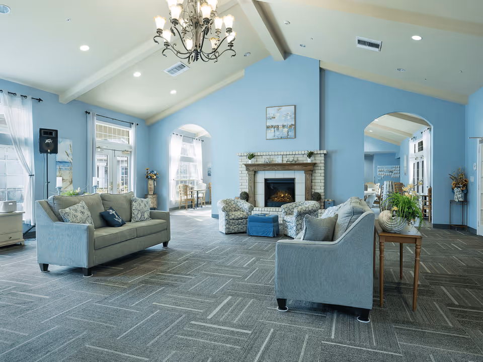 A spacious and well-lit living room area with light blue walls and a high ceiling. The room features two gray sofas with decorative pillows, two patterned armchairs, a blue ottoman, and a fireplace with a wooden mantel. There are large windows with sheer white curtains allowing natural light to enter. A chandelier hangs from the ceiling, and there are plants and decorative items on side tables. Arched doorways lead to adjacent rooms with more seating and tables.