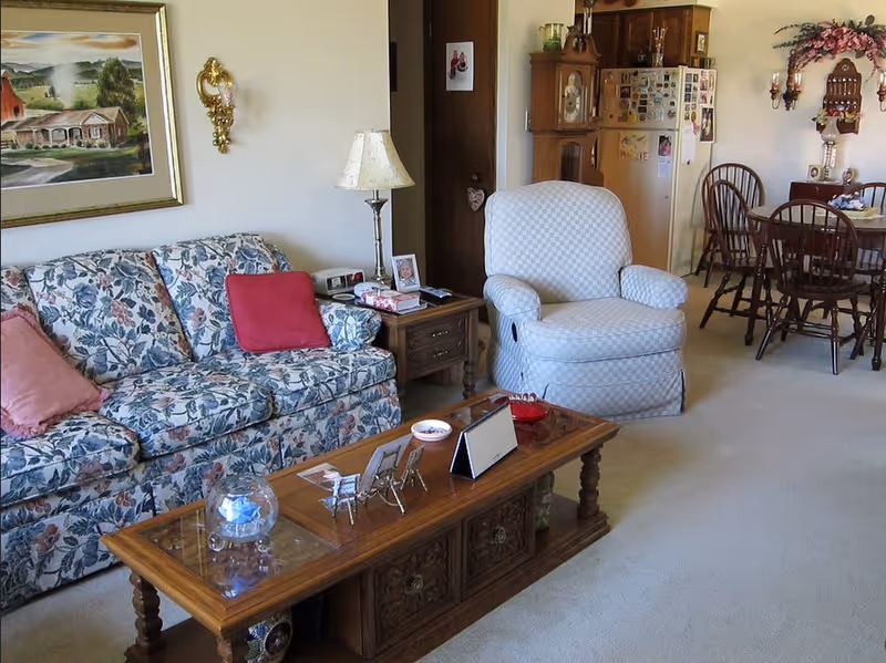 Cozy living room with a floral sofa, upholstered armchair, wooden coffee table and a dining area visible in the background.
