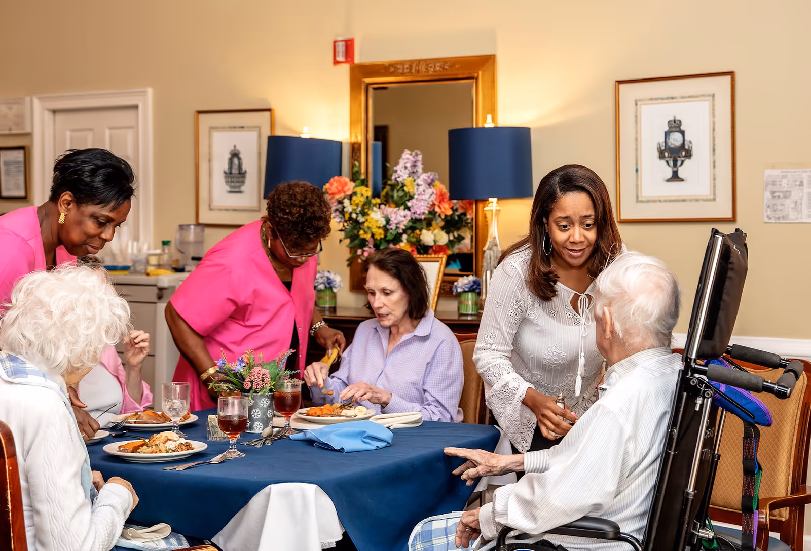 A group of elderly people and caregivers gathered around a dining table in a well-lit room. Two caregivers in pink uniforms are assisting the elderly individuals, while another woman in a white blouse is engaging with an elderly man in a wheelchair. The table is set with plates of food, glasses of iced tea, and a small flower arrangement. The background features a mirror, two blue lamps, and framed artwork on the walls.
