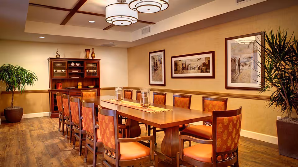 A well-lit dining room with a long wooden table surrounded by twelve chairs with orange patterned upholstery. The room features wooden flooring, two large potted plants, a wooden cabinet with dishes, and three framed artworks on the wall. Ceiling lights provide warm lighting.