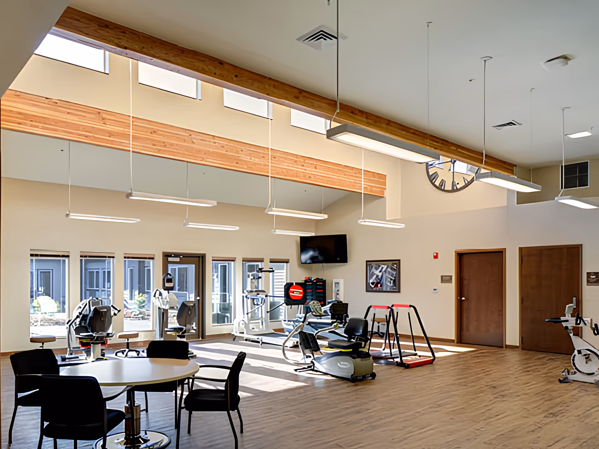 A spacious, well-lit exercise room with large windows and high ceilings featuring wooden beams. The room contains various exercise equipment including stationary bikes and other fitness machines. There is a round table with four chairs in the foreground and a wall-mounted TV on the far wall. The floor is wooden, and there are two closed wooden doors on the right side.