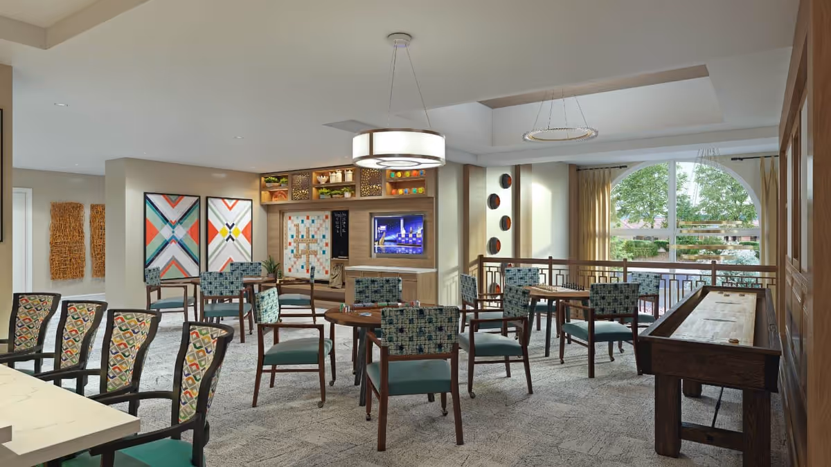 Bright community dining and activity room with multiple tables and patterned chairs, a shuffleboard table, TV and large arched window overlooking trees.