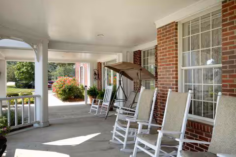 Covered front porch with white rocking chairs, a canopy swing, brick exterior and large windows overlooking landscaped grounds.