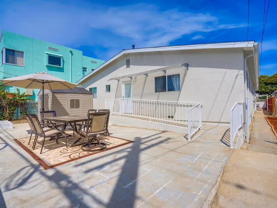 Outdoor patio area with a table and six chairs under a large umbrella on a decorative outdoor rug. There is a small storage shed next to the table. The patio is adjacent to a white building with a ramp and railing leading to a door and a window with an awning. The sky is clear and blue.