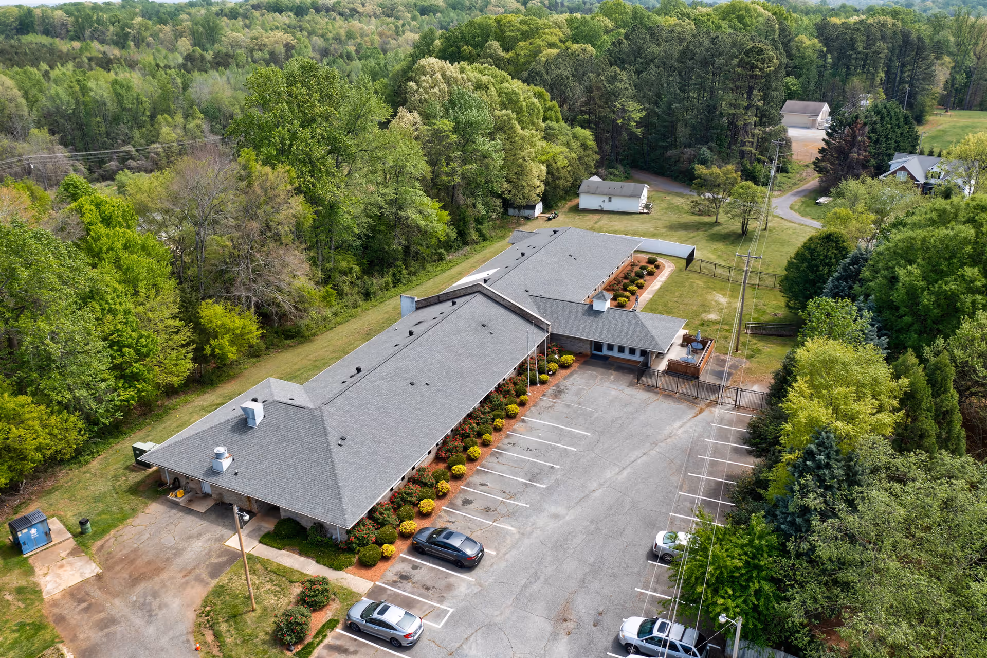 Aerial view of a single-story building with a gray roof surrounded by greenery and trees. The building is adjacent to a parking lot with several parked cars. There are small bushes and landscaped areas along the building's perimeter. The setting appears to be in a rural or suburban area with a mix of open grassy spaces and dense trees.