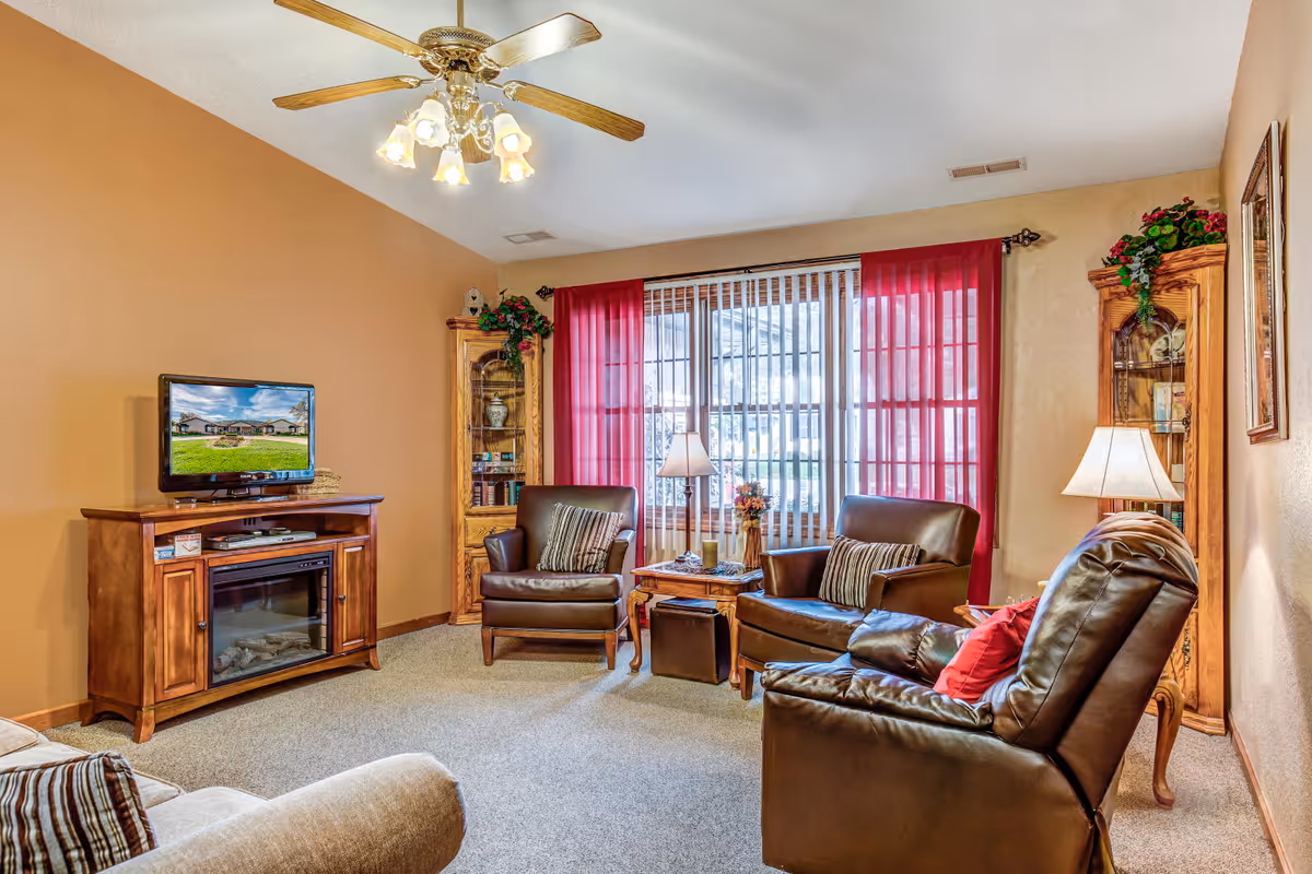 A cozy living room with beige walls and carpeted floor. The room features a ceiling fan with lights, a wooden TV stand with a flat-screen TV, two wooden display cabinets with glass doors, and three leather armchairs arranged around a wooden coffee table. The window is covered with vertical blinds and red curtains, and there are two table lamps providing warm lighting.
