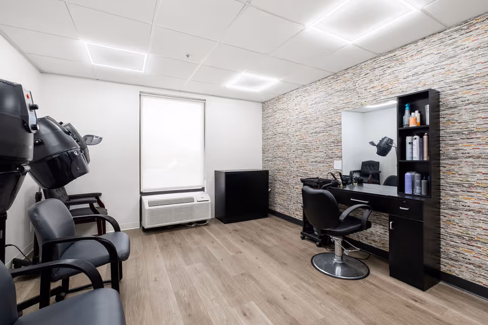 A small salon room with three black salon chairs, two hair dryers mounted on the left wall, a large window with a white blind, a black cabinet, and a black styling station with a mirror and shelves holding hair care products. The room has light wood flooring and a textured accent wall behind the styling station.