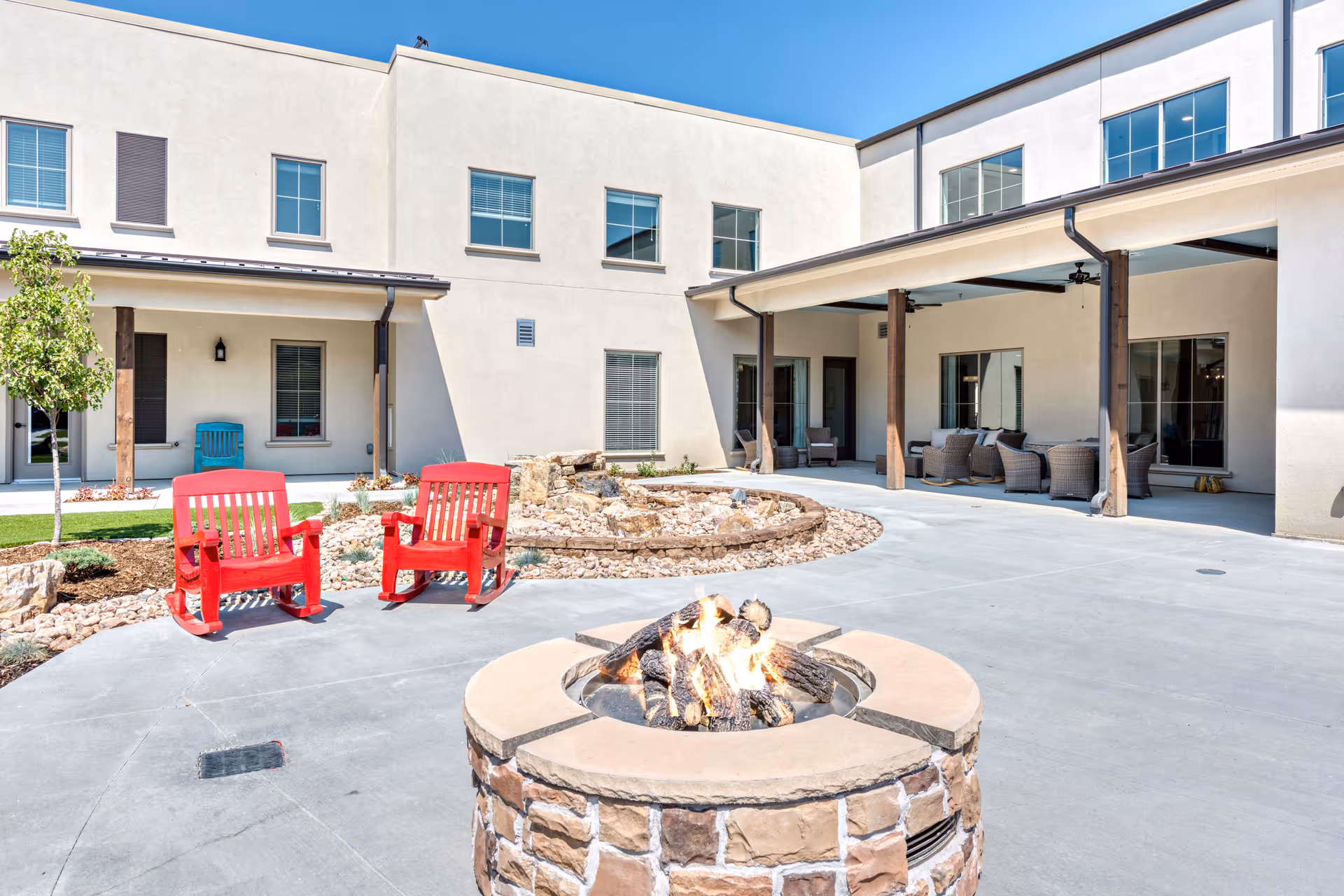 Outdoor courtyard area of a senior living facility featuring a stone fire pit with a burning fire in the foreground, two bright red rocking chairs, a small landscaped garden with rocks and plants, and a covered patio with wicker seating. The building has beige walls and multiple windows under a clear blue sky.