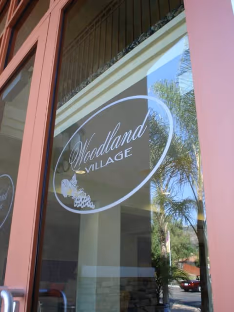 Exterior entrance door with Woodland Village sign reflected in glass window, palm trees nearby.